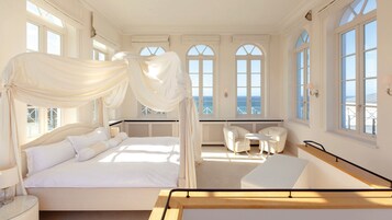 Quarto com cama de dossel, área de estar com mesa e cadeiras, janelas amplas com vista para o oceano e uma varanda.