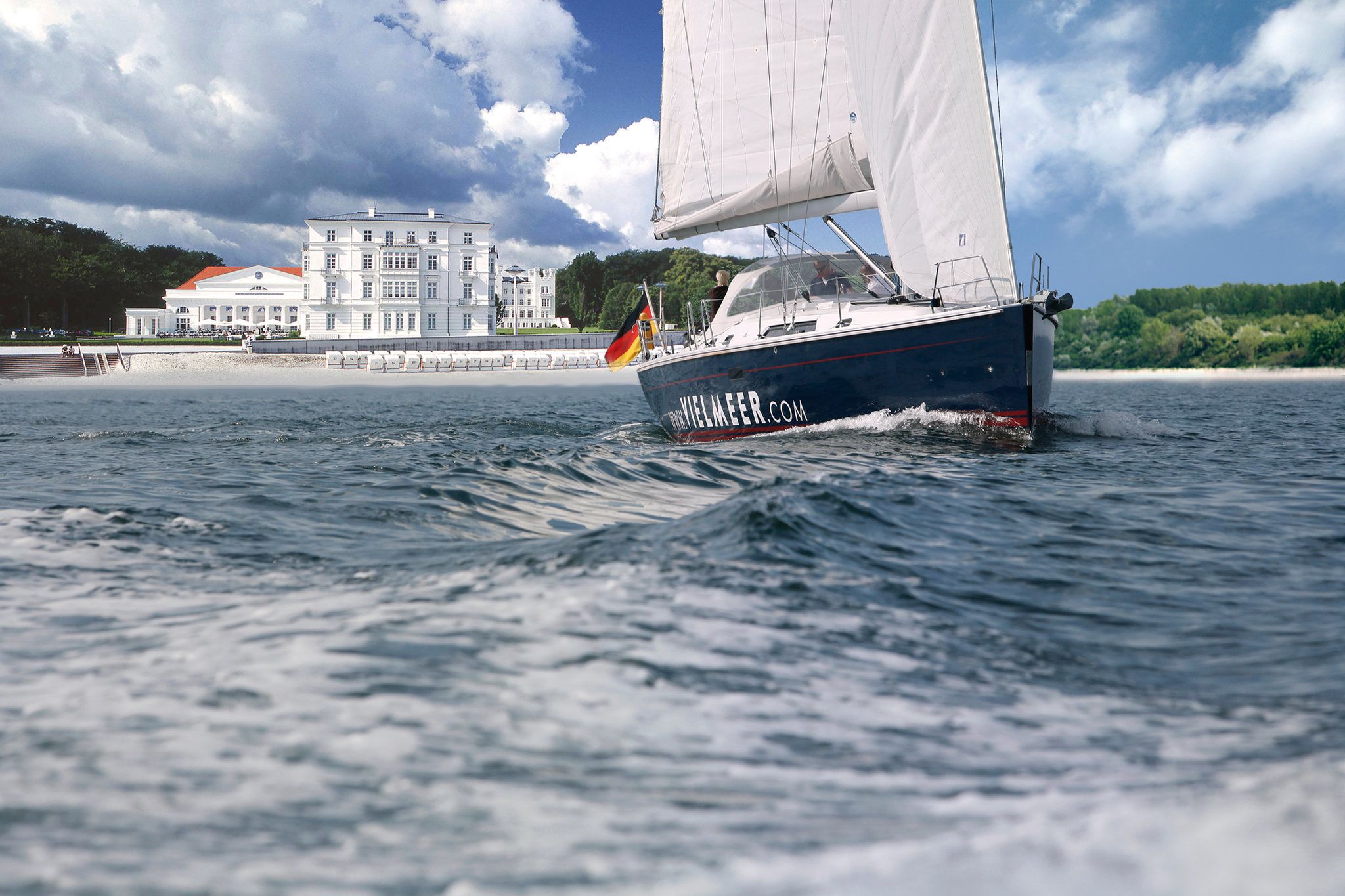 A sailboat with a German flag on the water, a large building with a red roof in the background, and a beach with steps leading down to the water.