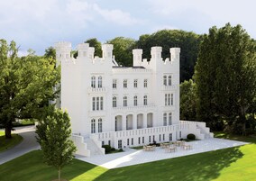 Um edifício grande e branco, semelhante a um castelo, com diversas torres e janelas em arco, cercado por vegetação exuberante.