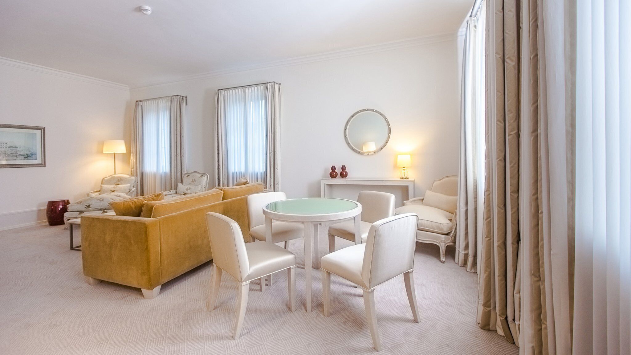 Een hotelkamer met een bank, stoelen, een ronde tafel, een spiegel en gordijnen.