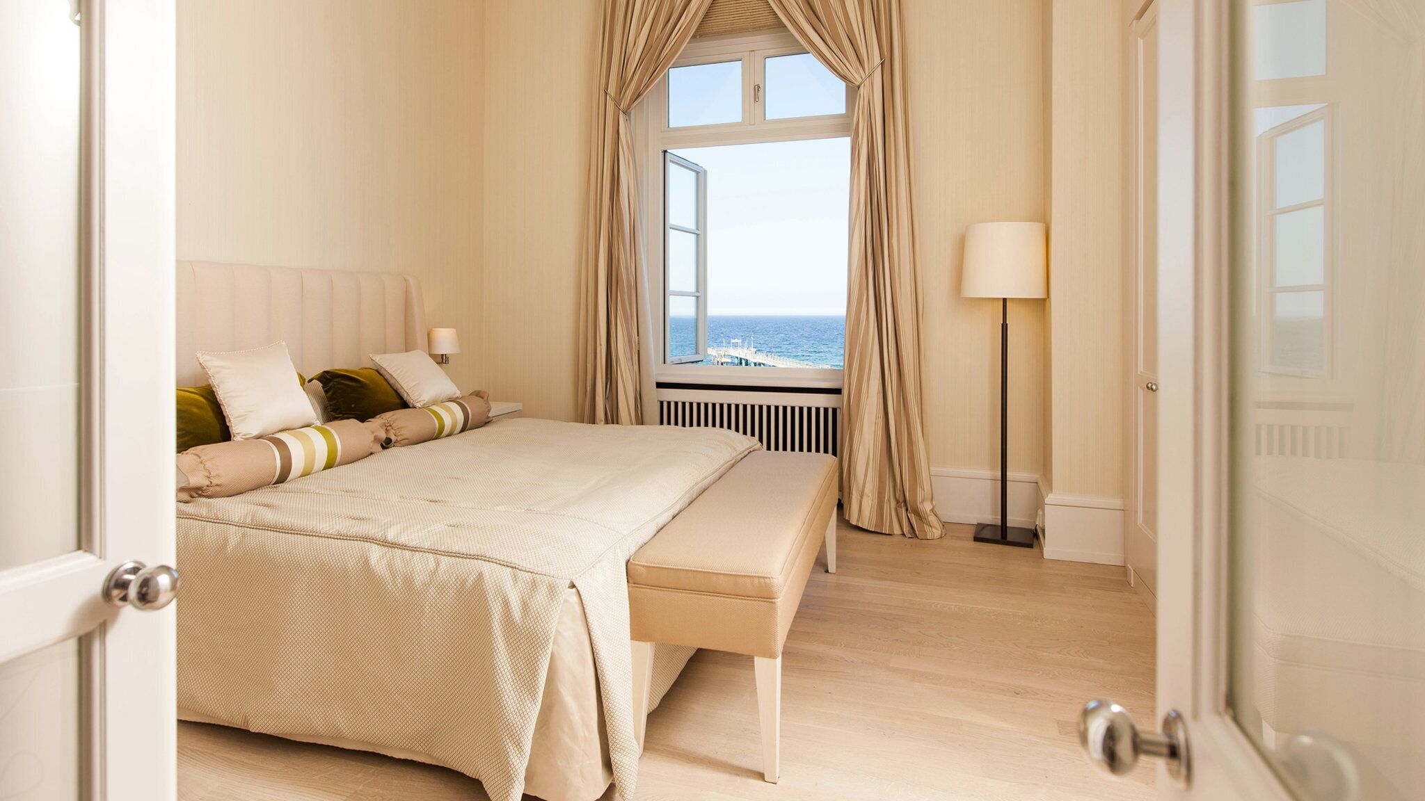 Un dormitorio con una cama grande, una banca, una mesita de noche, una lámpara y vistas al mar a través de una ventana.