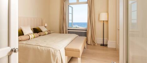 Un dormitorio con una cama grande, una banca, una mesita de noche, una lámpara y vistas al mar a través de una ventana.