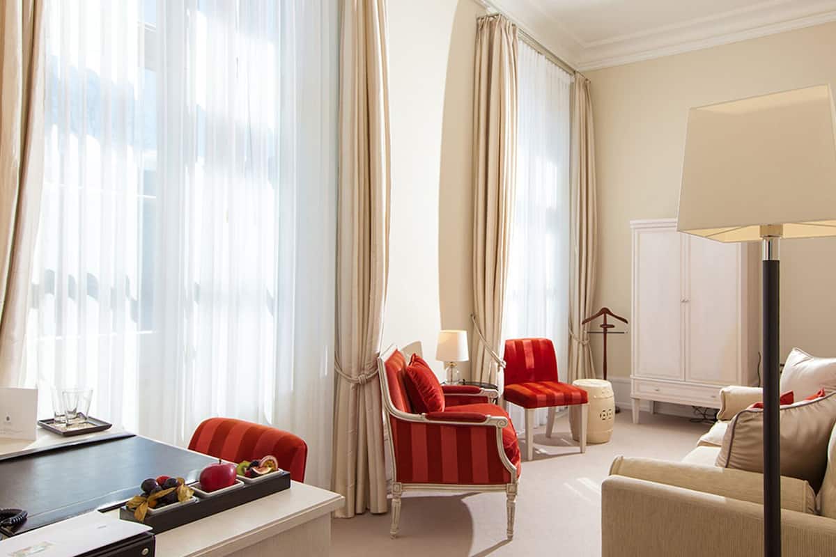 Une chambre d’hôtel avec un bureau, deux chaises rouges, un canapé beige, une lampe sur pied et une armoire.
