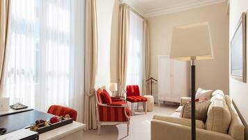 Habitación de hotel con escritorio, dos sillas rojas, un sofá color beige, una lámpara de pie y un armario.