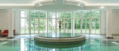 A large indoor swimming pool with a central fountain, surrounded by large windows offering views of greenery.