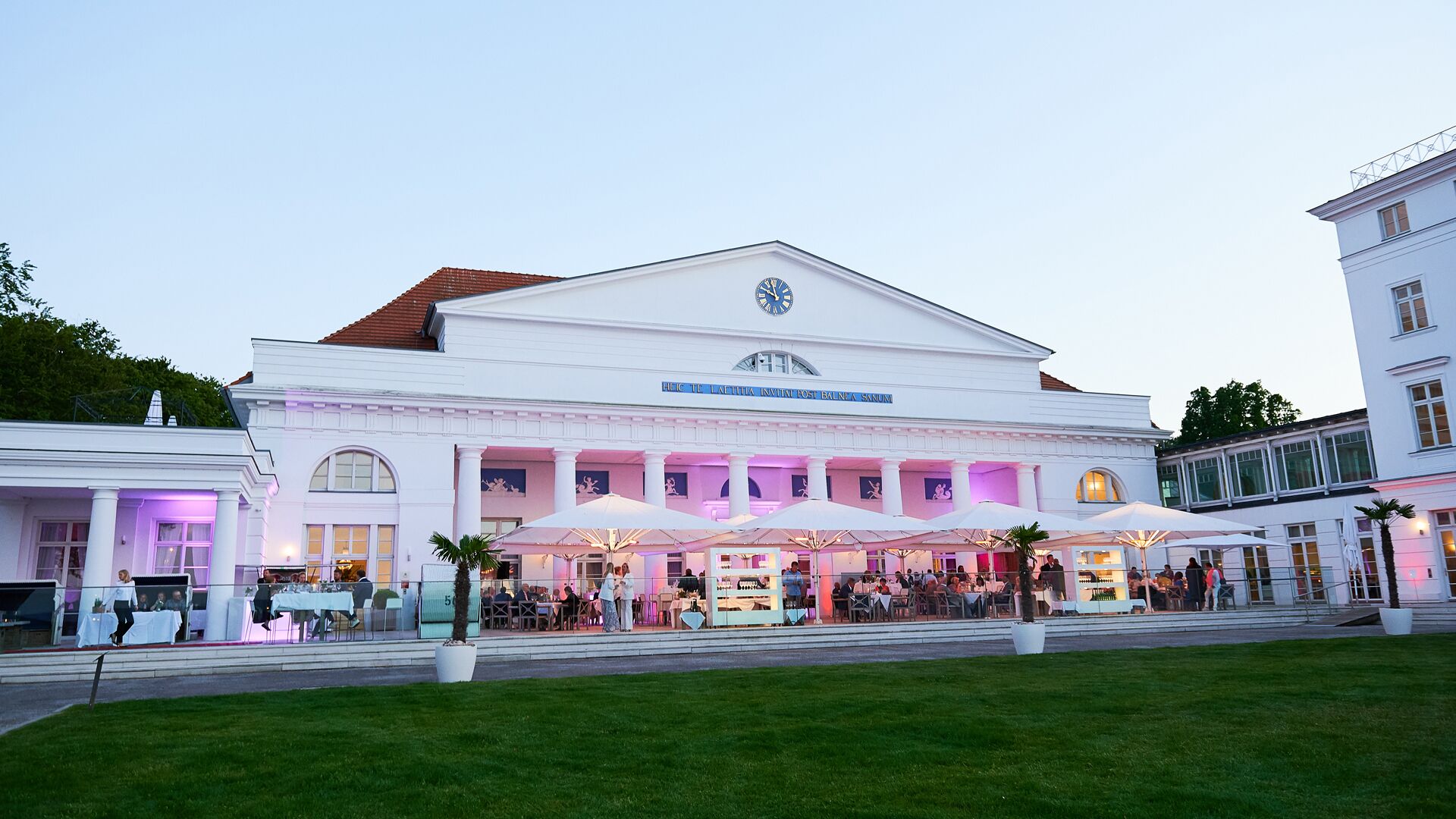 A historic building with a clock tower, large white facade, and outdoor seating area with umbrellas.