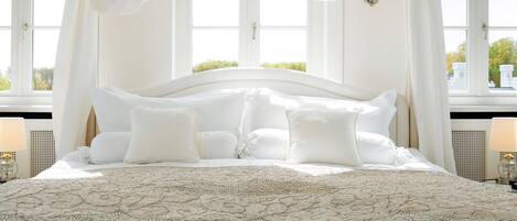 A large bed with a canopy, multiple pillows, and a patterned bedspread in a room with large windows and a view of greenery outside.