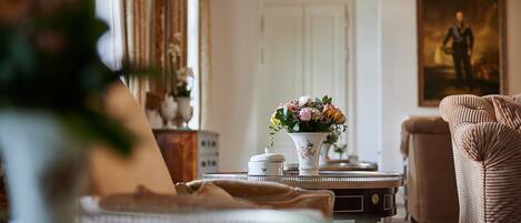 A well-lit room with a classic interior design, featuring a white table with a vase of flowers, a patterned armchair, and a framed portrait on the wall.