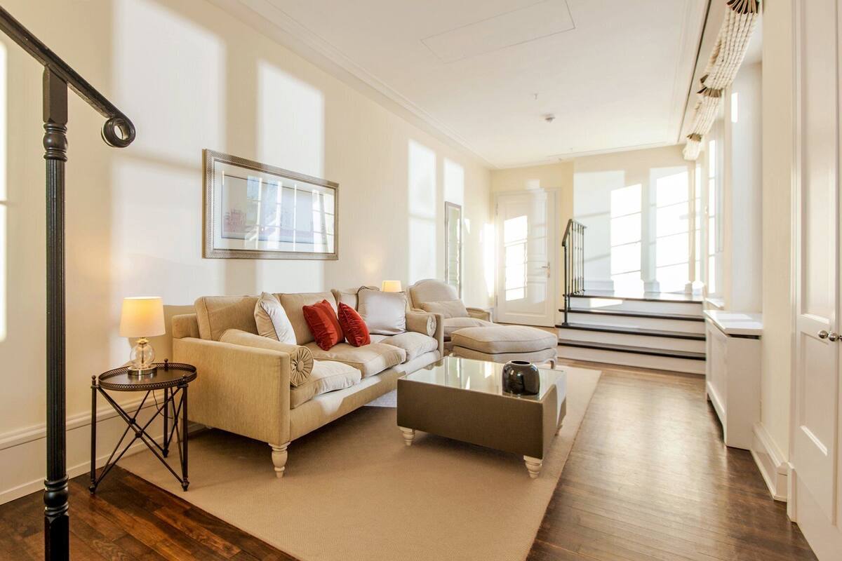 Un salon spacieux avec un canapé beige, une table basse et une table d’appoint avec une lampe. Un escalier mène à un autre niveau.