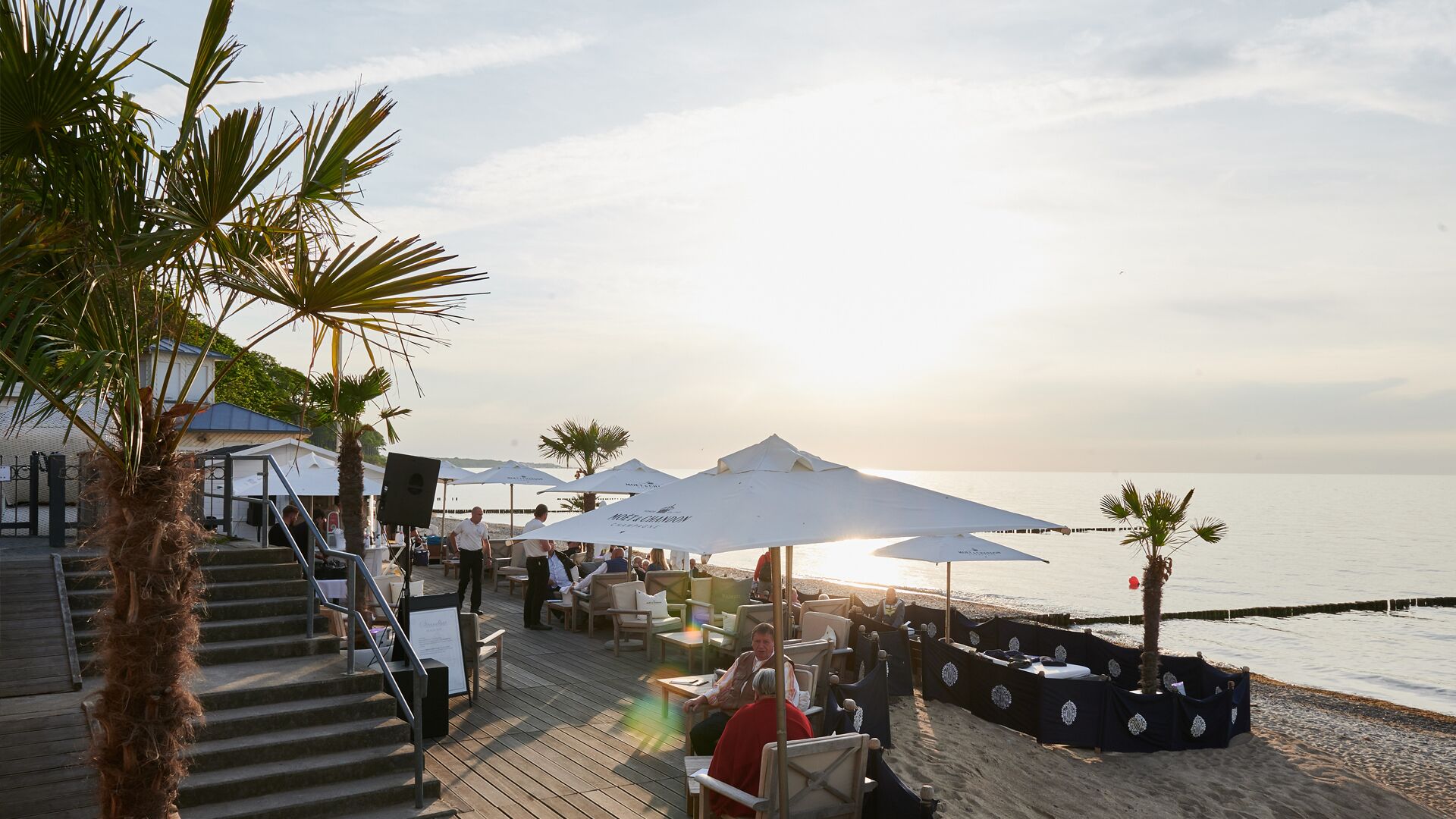 A beachside restaurant with outdoor seating, umbrellas, and a view of the sea.