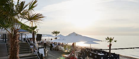 A beachside restaurant with outdoor seating, umbrellas, and a view of the sea.