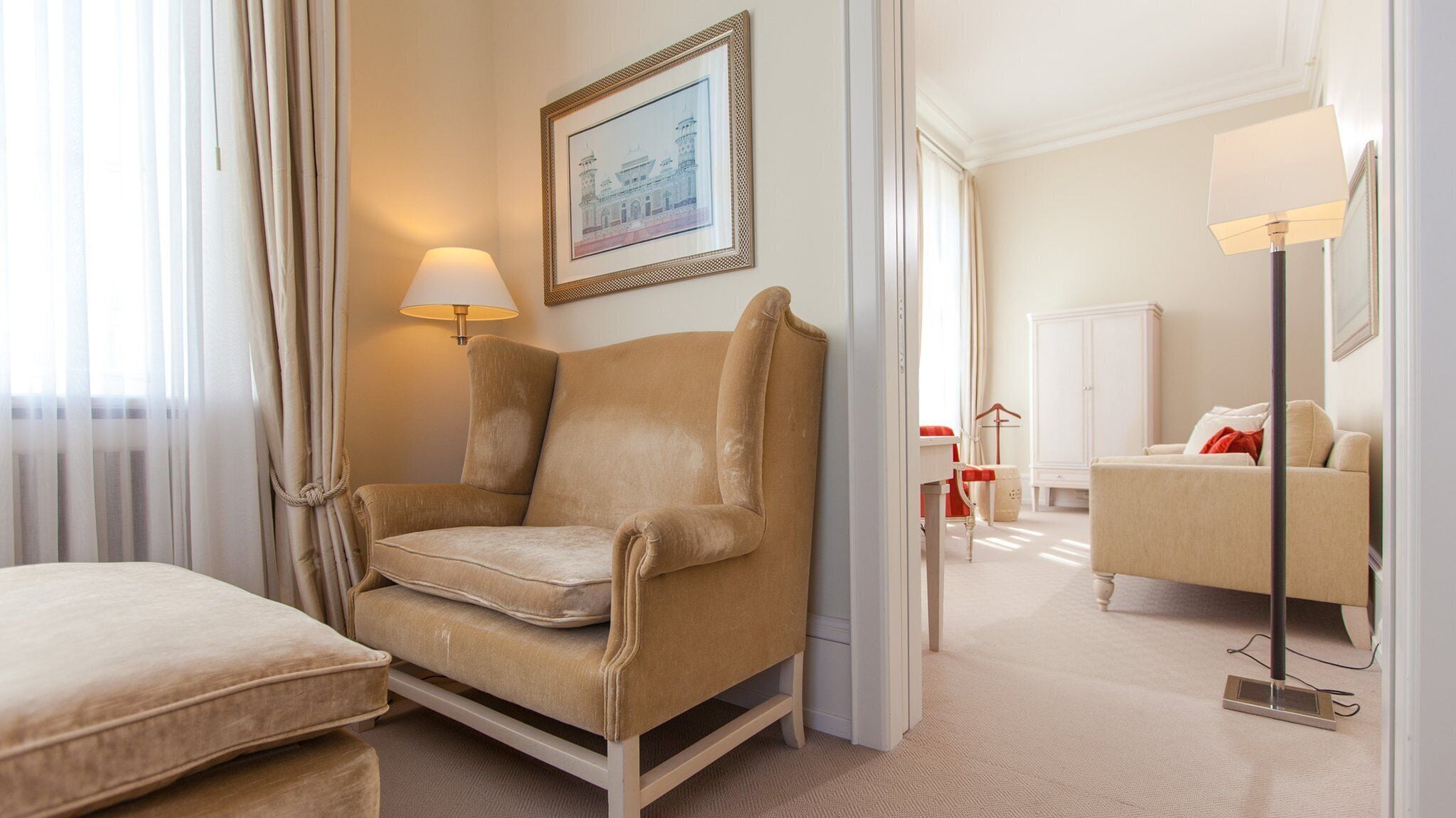A hotel room with a beige armchair, a small table, a lamp, a framed picture on the wall, and a sofa in the background.