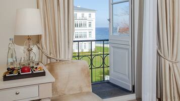 A hotel room with a balcony, a table with a lamp and fruit, and a view of a white building across the water.