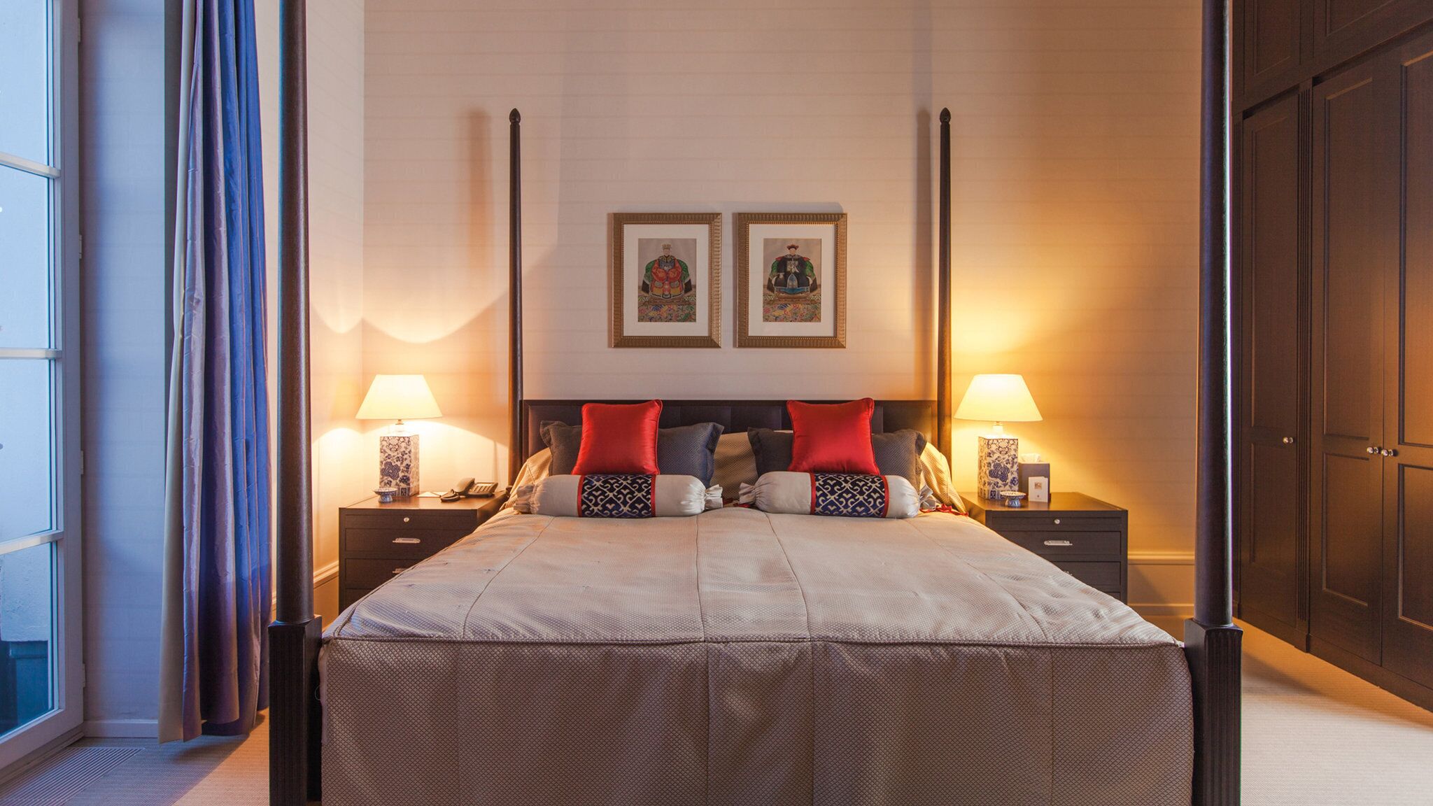 A four-poster bed with red and patterned pillows, two framed artworks above, and bedside lamps on either side.