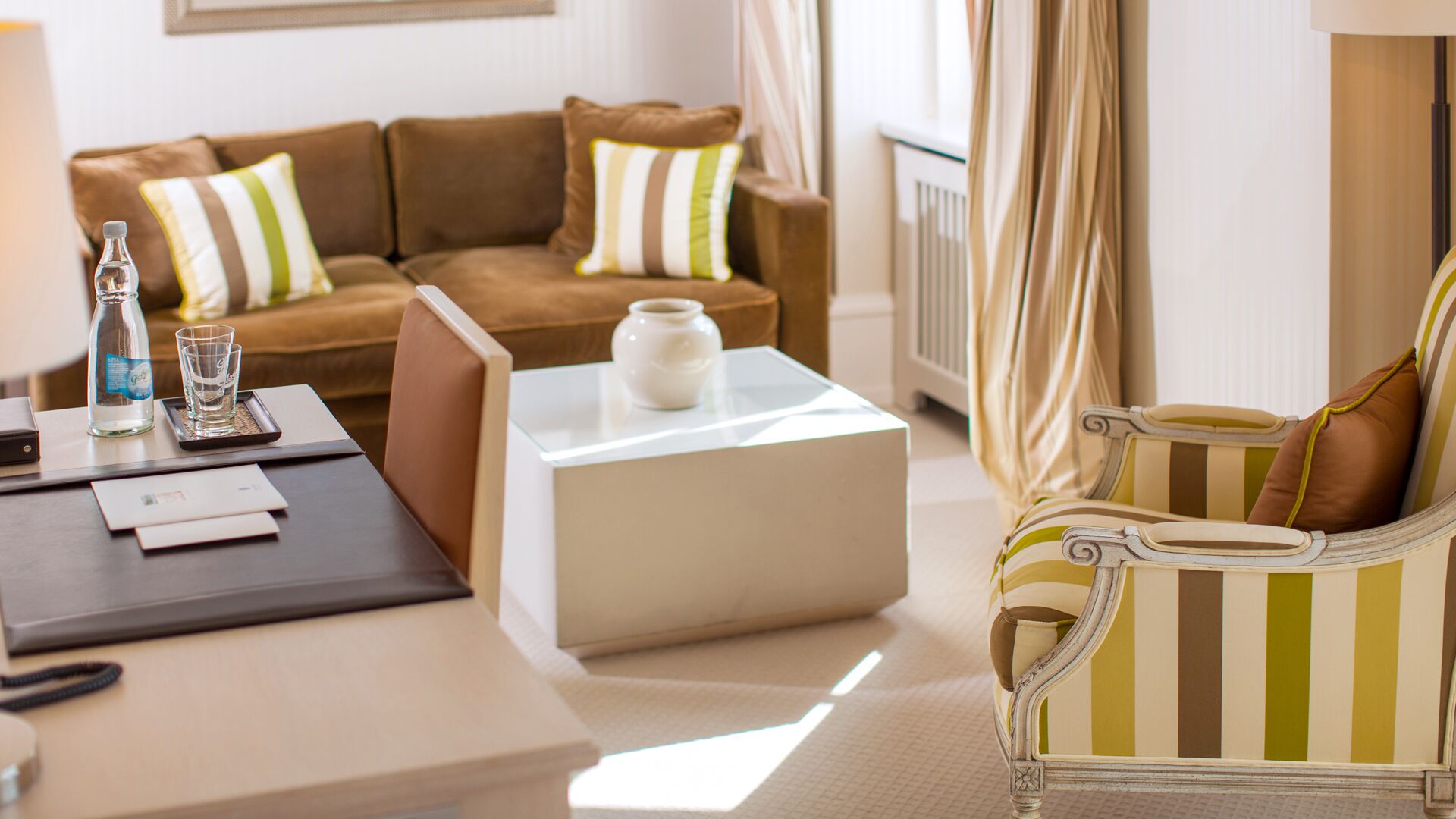 A hotel room with a brown sofa, a striped armchair, a white coffee table, and a small white vase.