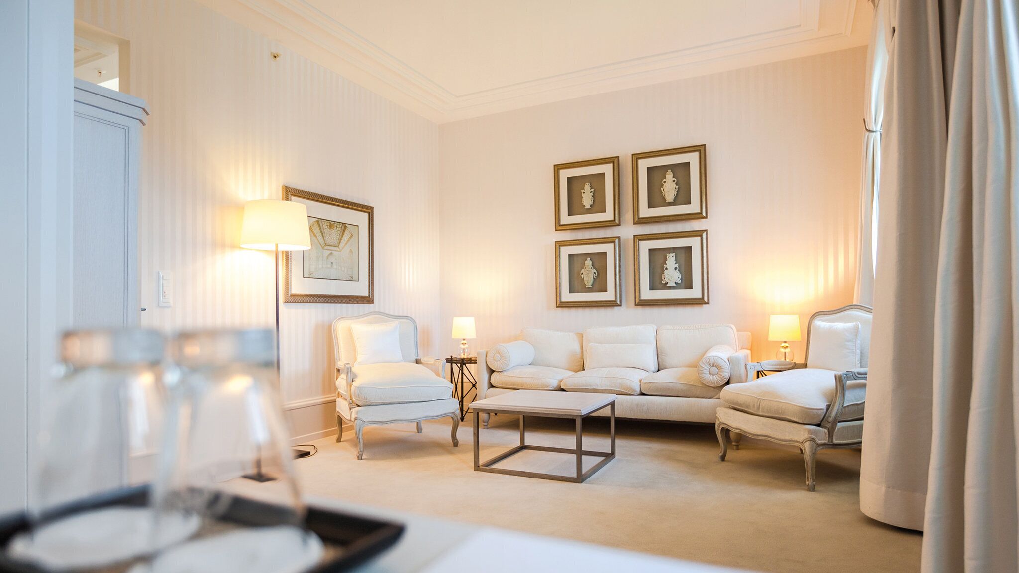 A spacious living room with a white sofa set, a coffee table, and a side table with a lamp. There are framed pictures on the wall and a door with a light switch visible.