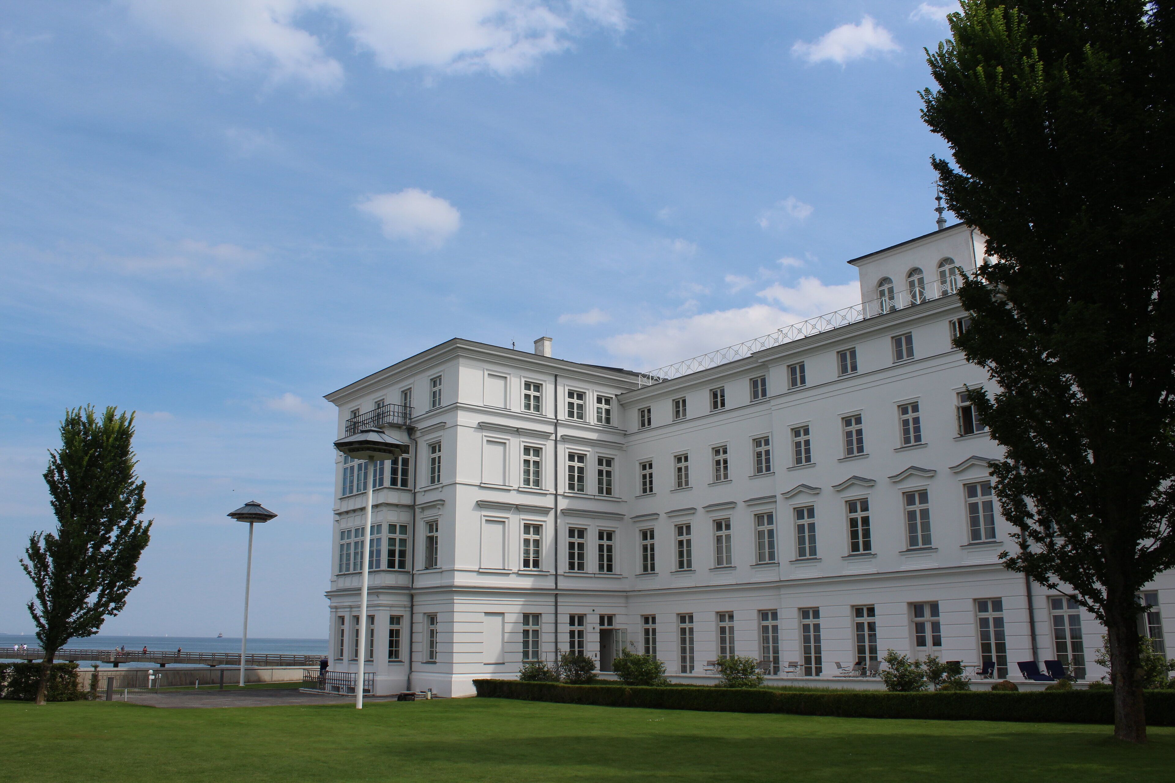 A large, white building with multiple windows and a balcony, surrounded by well-maintained lawns and trees.