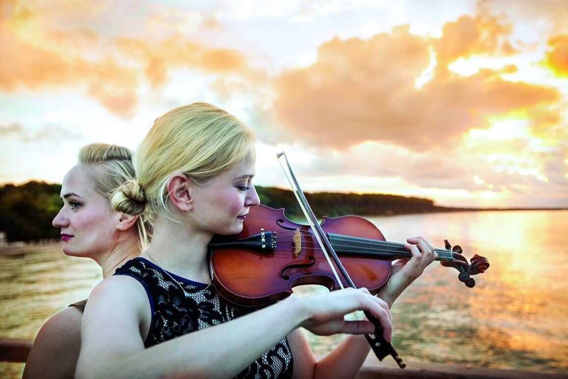 Dos personas tocan violines junto a un cuerpo de agua al atardecer.