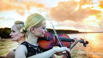 Dos personas tocan violines junto a un cuerpo de agua al atardecer.