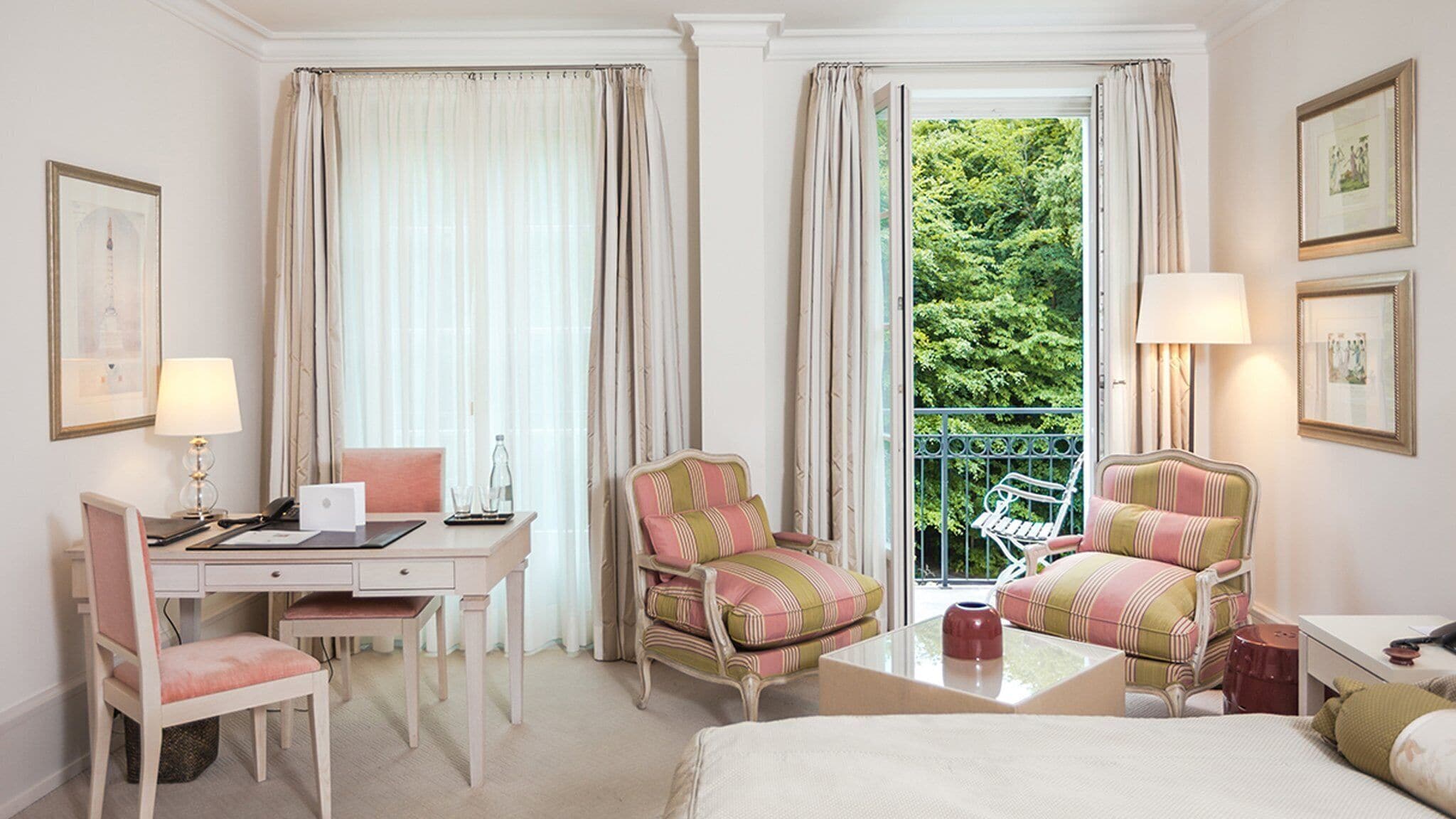 Habitación de hotel con una cama, dos sillones, un escritorio con una computadora portátil, una silla, una mesita y un balcón con vistas a árboles.