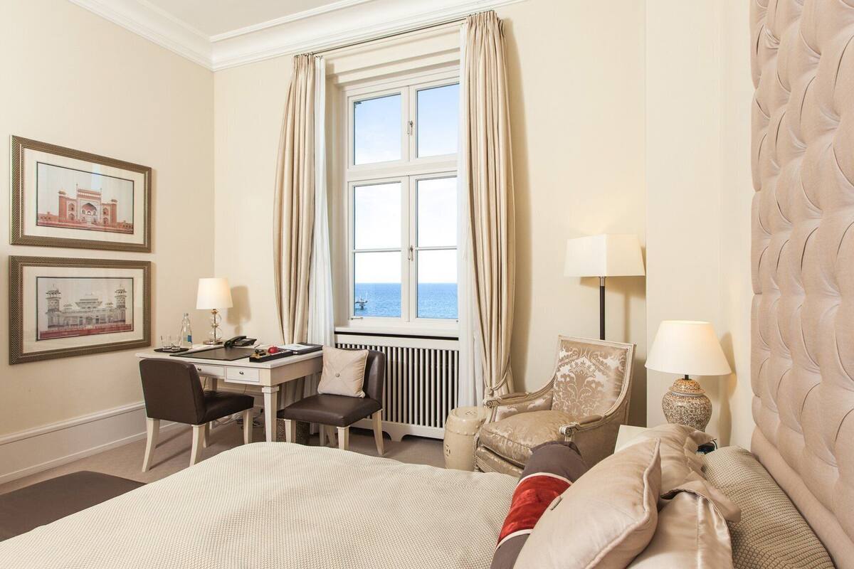 Une chambre d’hôtel avec un grand lit, un bureau et une chaise. Il y a deux lampes sur le bureau et un radiateur en dessous.