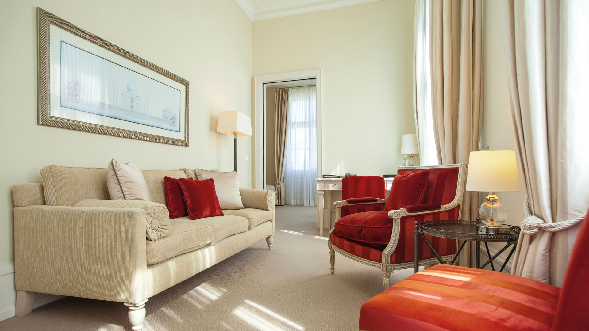 A hotel room with a beige sofa, a red armchair, a small round table with a lamp, and a framed picture on the wall.