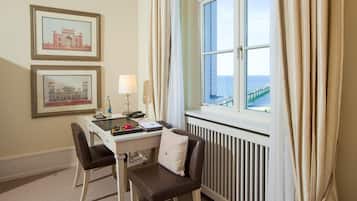Ein Hotelzimmer mit Schreibtisch, zwei Stühlen, einer Heizung, einem Fenster mit Vorhängen und zwei gerahmten Bildern an der Wand.