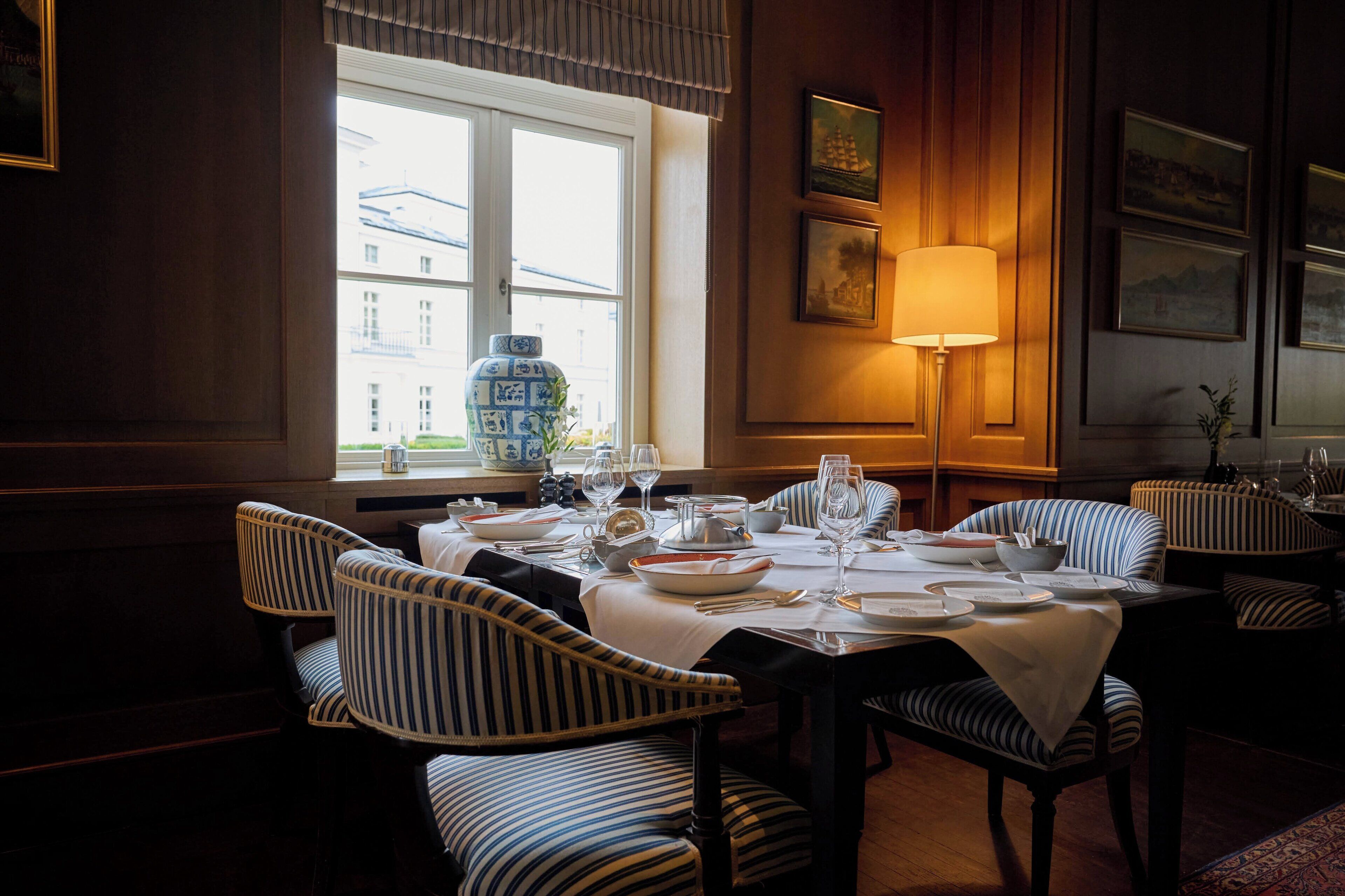 Un coin repas avec des chaises à rayures et une table dressée pour le repas, un vase sur le rebord de la fenêtre et des tableaux encadrés au mur.