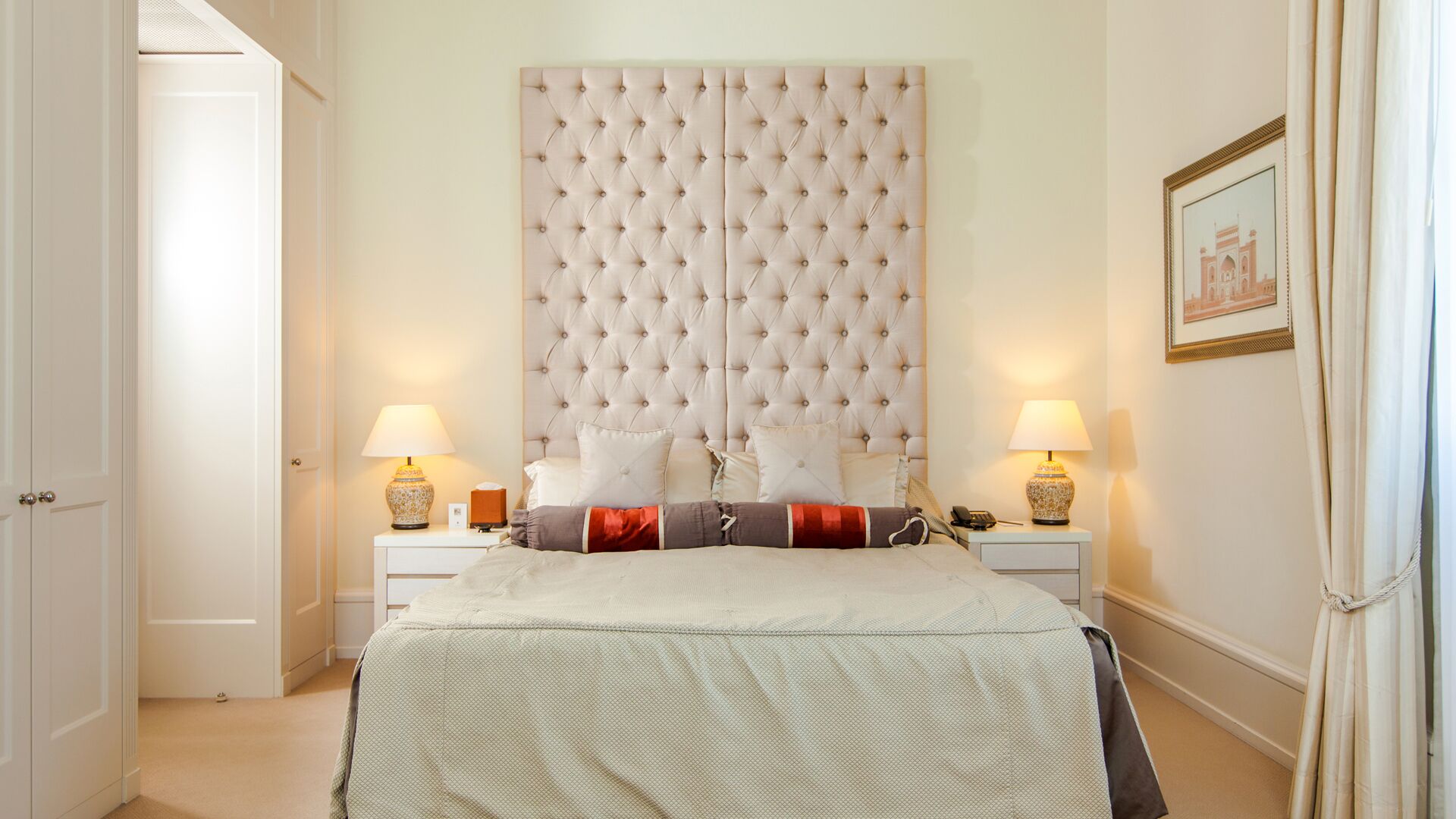 Een slaapkamer met een geborduurde hoofdbank, twee nachtkastjes met lampen, een bed met een beddengoed met patroon, en een ingelijst schilderij aan de muur.