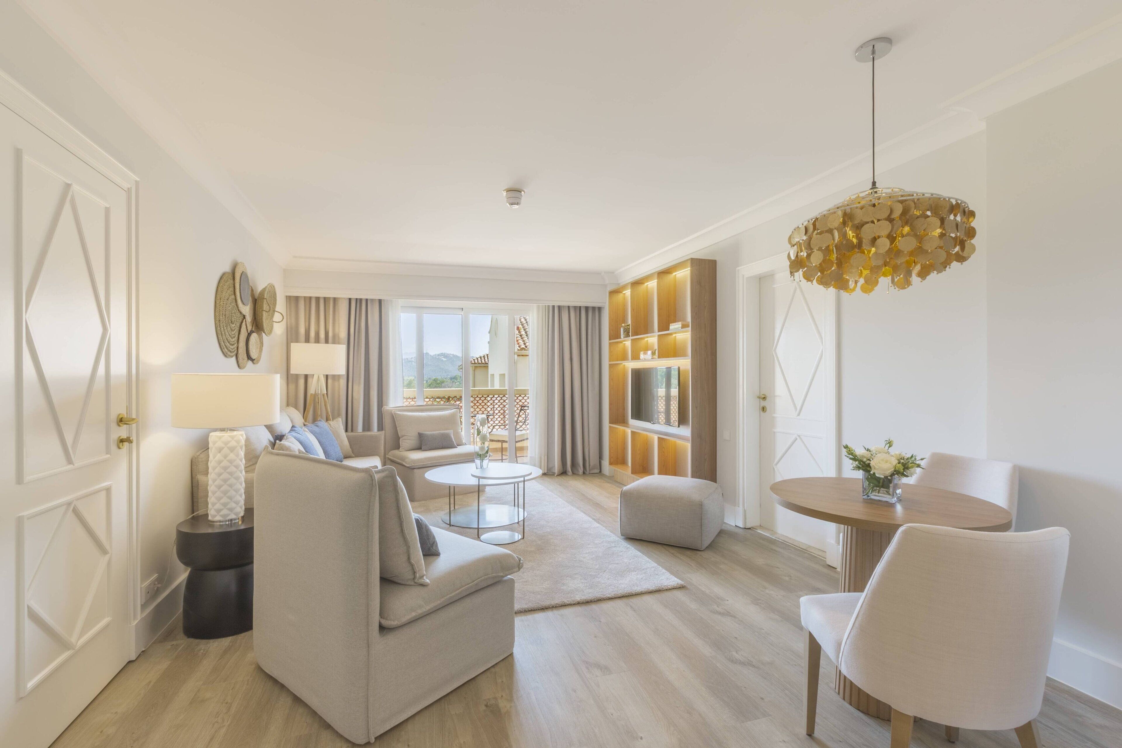 Een moderne woonkamer met een bank, fauteuils, een salontafel en een eethoek met stoelen en een tafel. Er is een groot raam met gordijnen, een wandtelevisie en een decoratieve lamp.