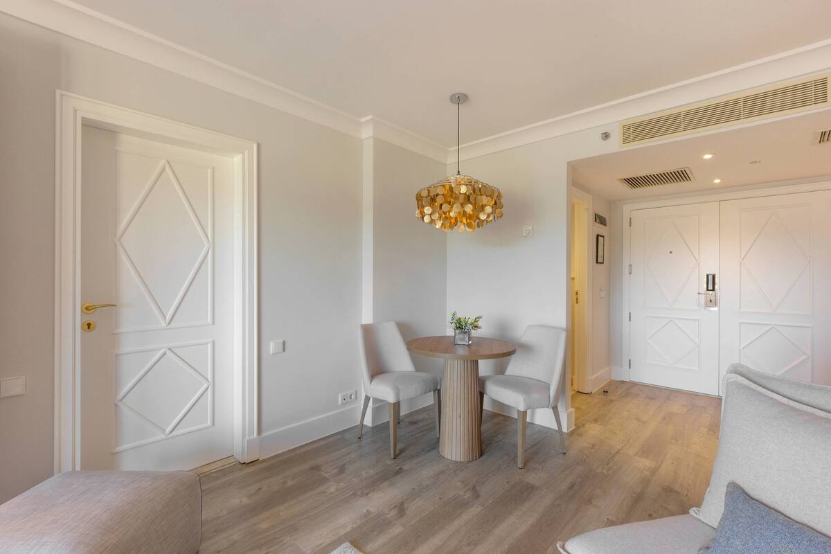 Quarto moderno com mesa de jantar redonda, duas cadeiras, luminária pendente e porta branca com padrão de losangos.