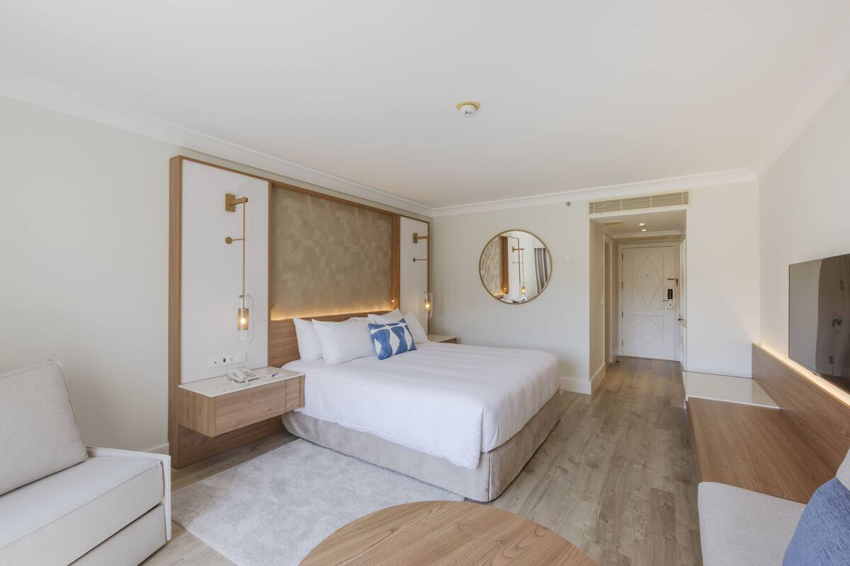 Quarto de hotel moderno com uma cama grande, mesas de cabeceira, um espelho redondo e uma televisão.
