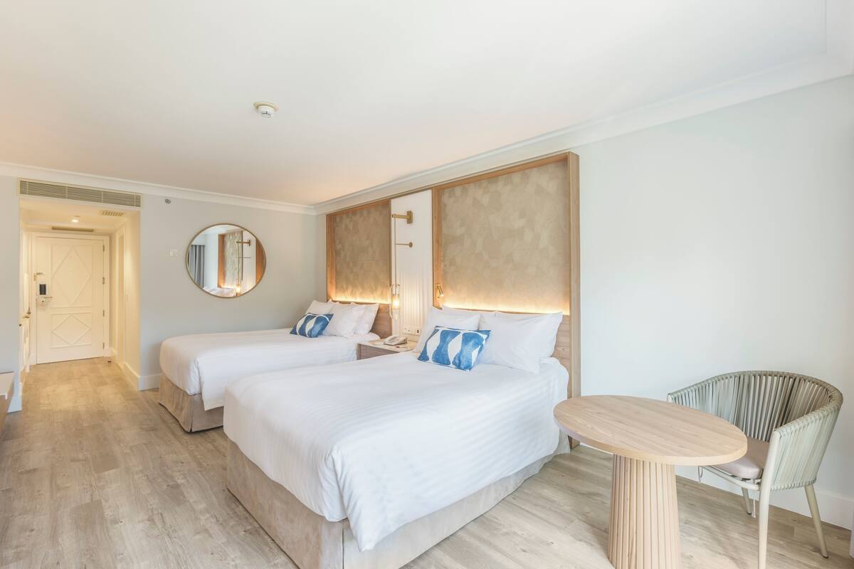 Quarto de hotel com duas camas, um espelho redondo, uma mesinha e uma cadeira de vime.