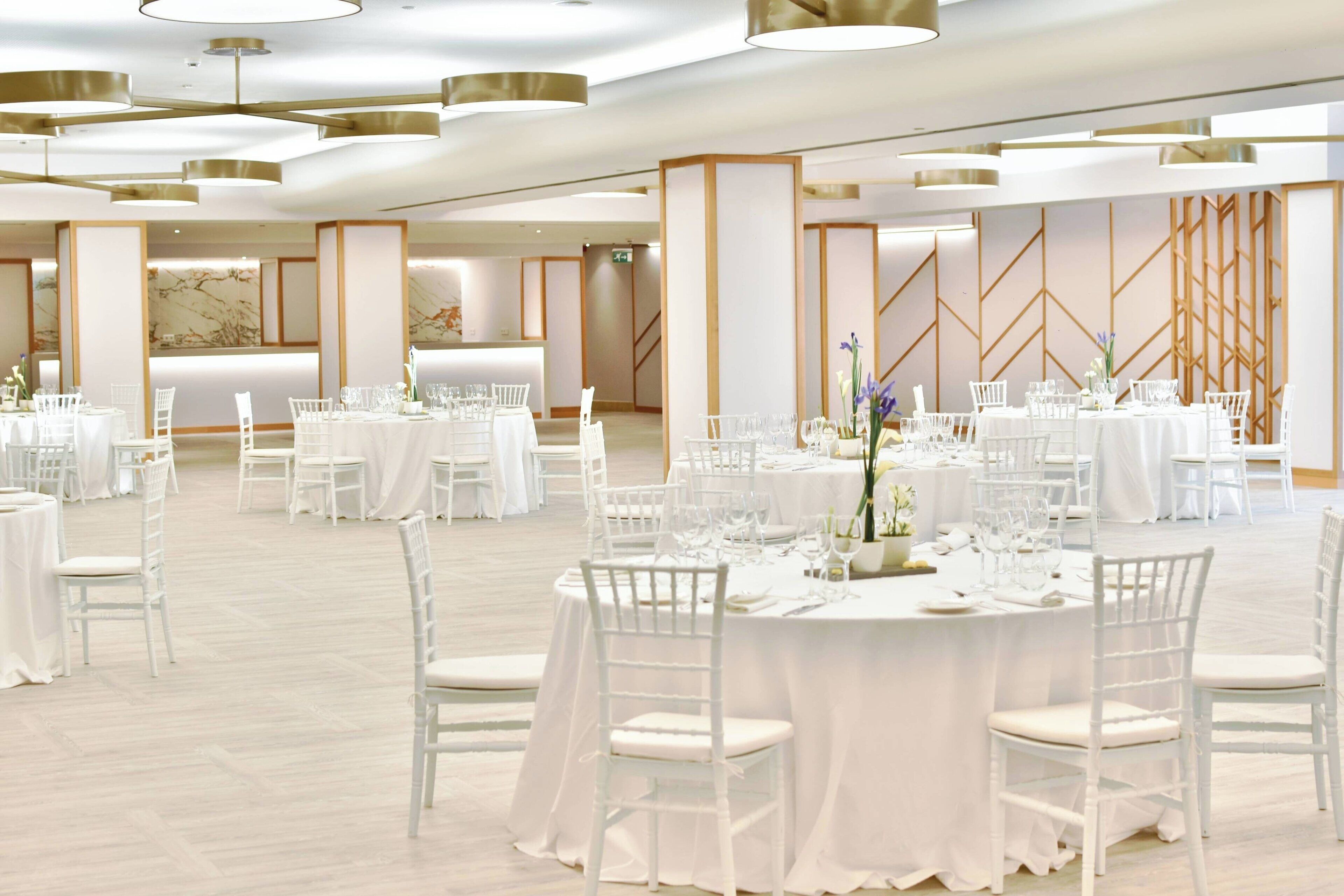 Un amplio salón de banquetes con mesas redondas dispuestas para comer, sillas blancas y modernos candelabros.