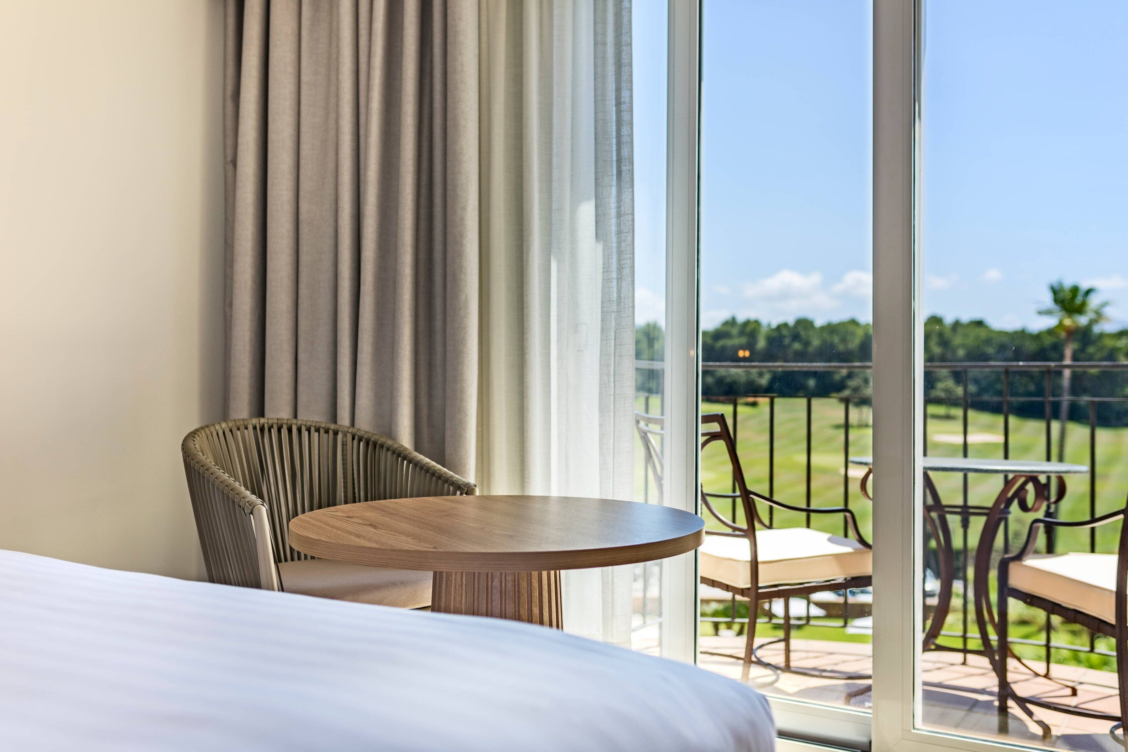 Habitación de hotel con una cama, una mesa redonda de madera y una silla de mimbre. Hay un balcón con vistas a un campo de golf.