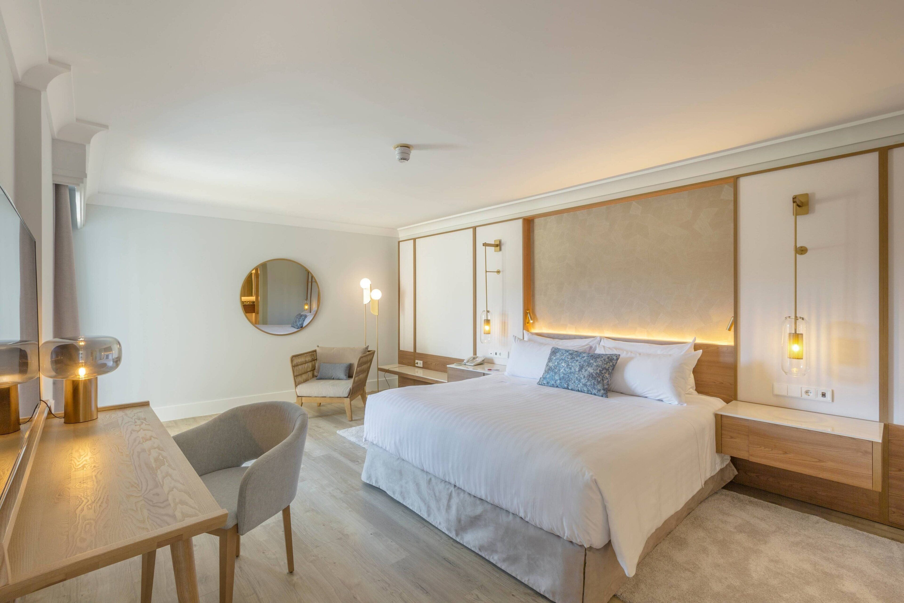 Una camera d'albergo moderna con un letto grande, una scrivania con una sedia, uno specchio rotondo e un comodino con una lampada.
