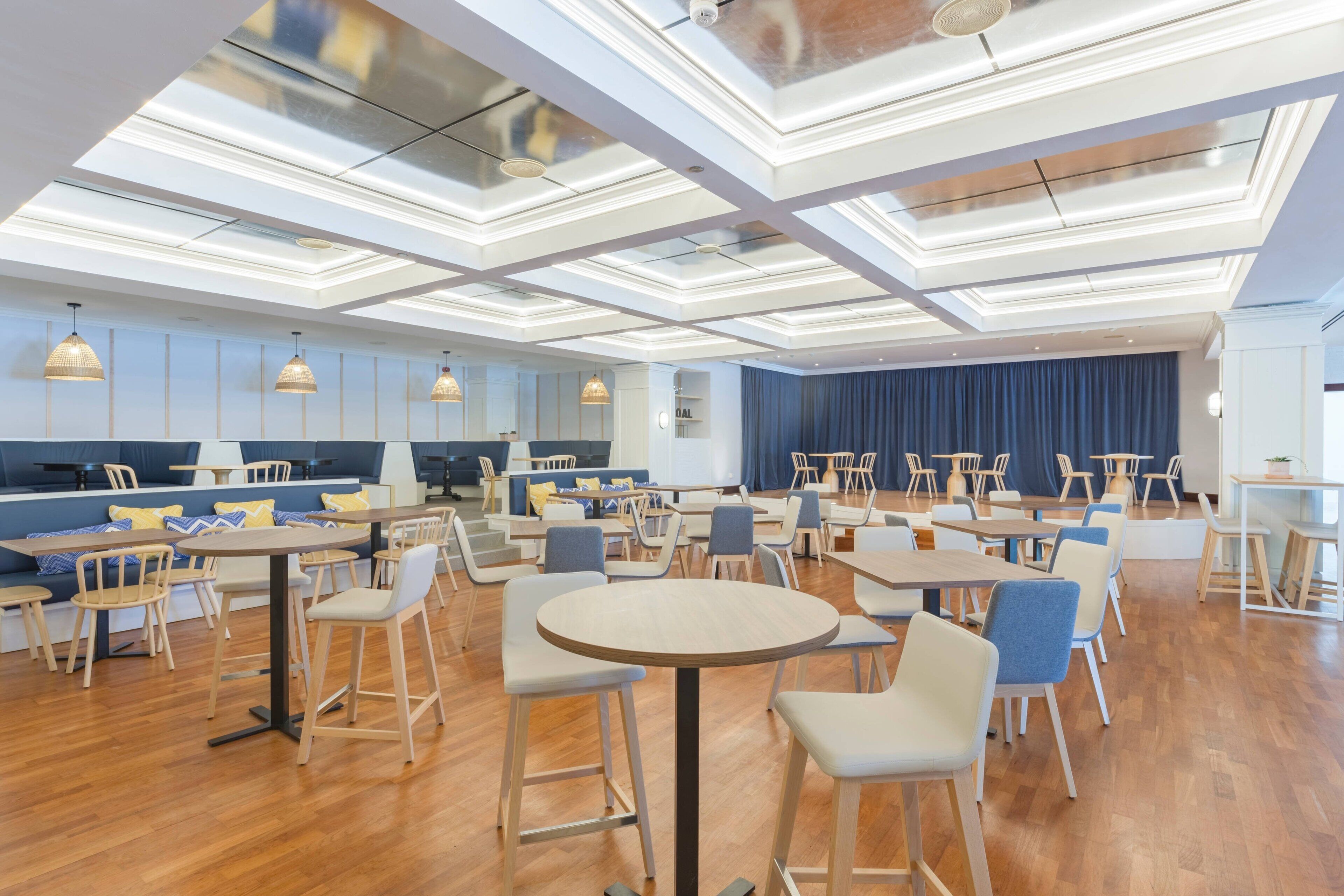 A spacious dining area with multiple tables and chairs, a high ceiling with recessed lighting, and a blue curtain backdrop.