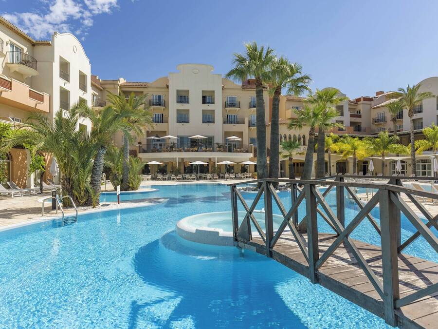 Piscina de un resort con muelle de madera, palmeras y un edificio con balcones y terraza.