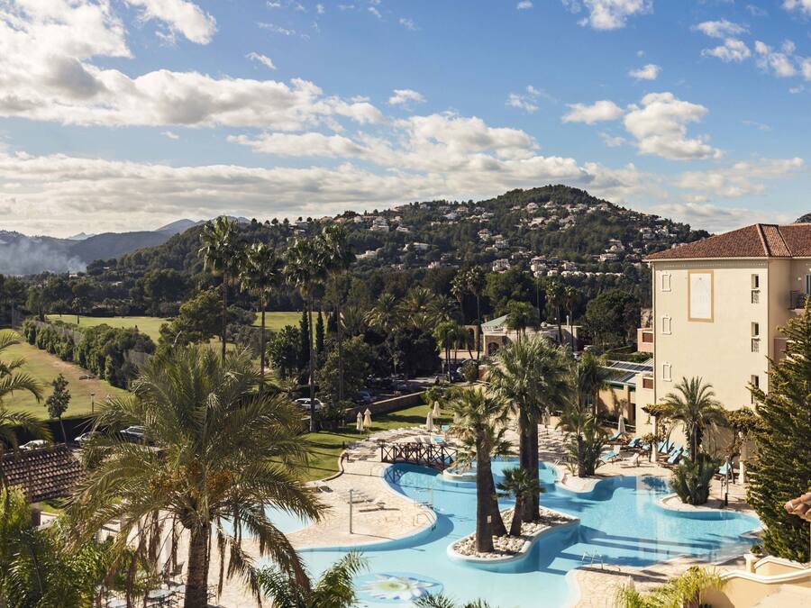 A resort with a swimming pool, palm trees, and a view of a hilly landscape.