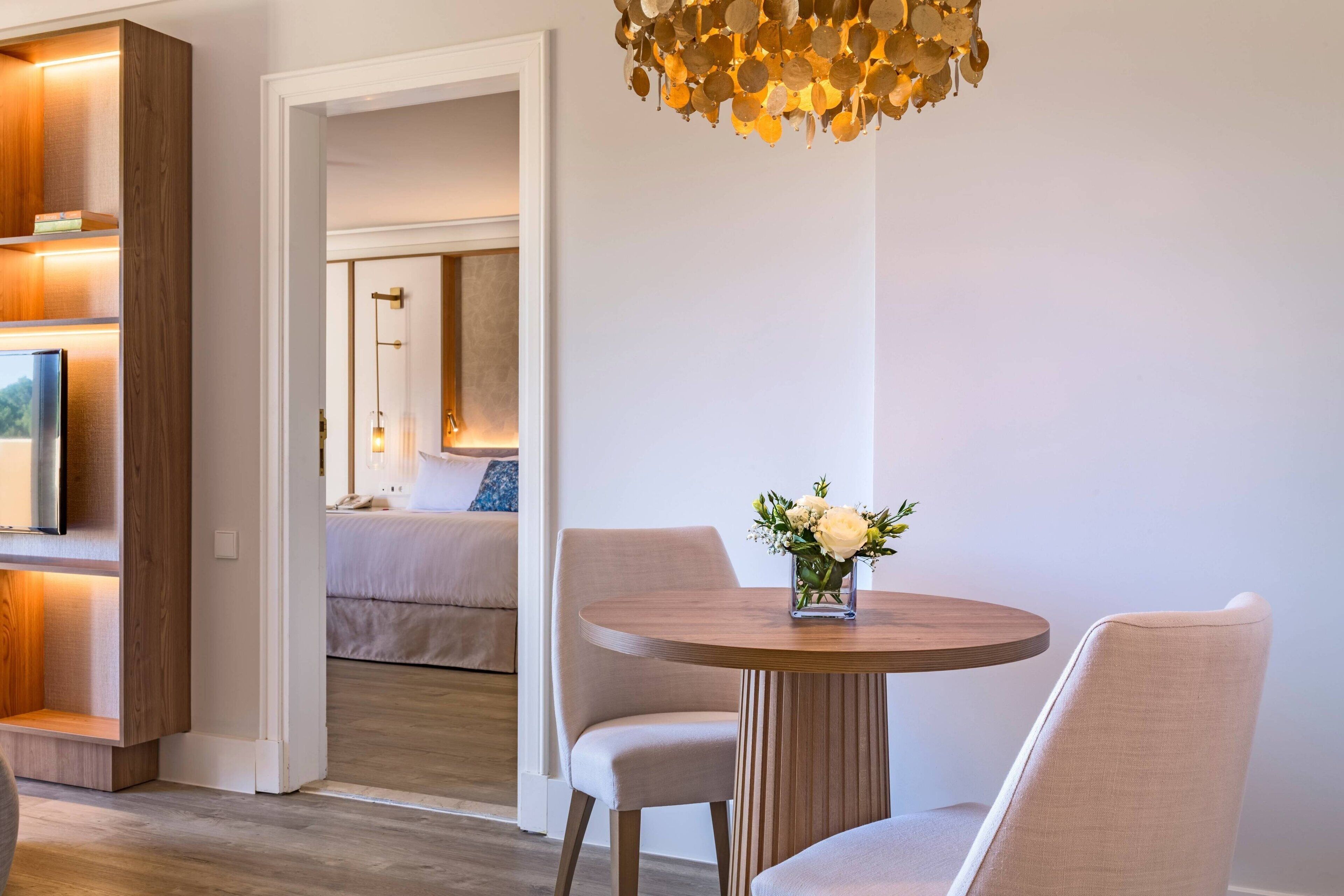 Habitación de hotel moderna con piso de madera, una mesa redonda con sillas, un jarrón de flores y una lámpara de araña original.