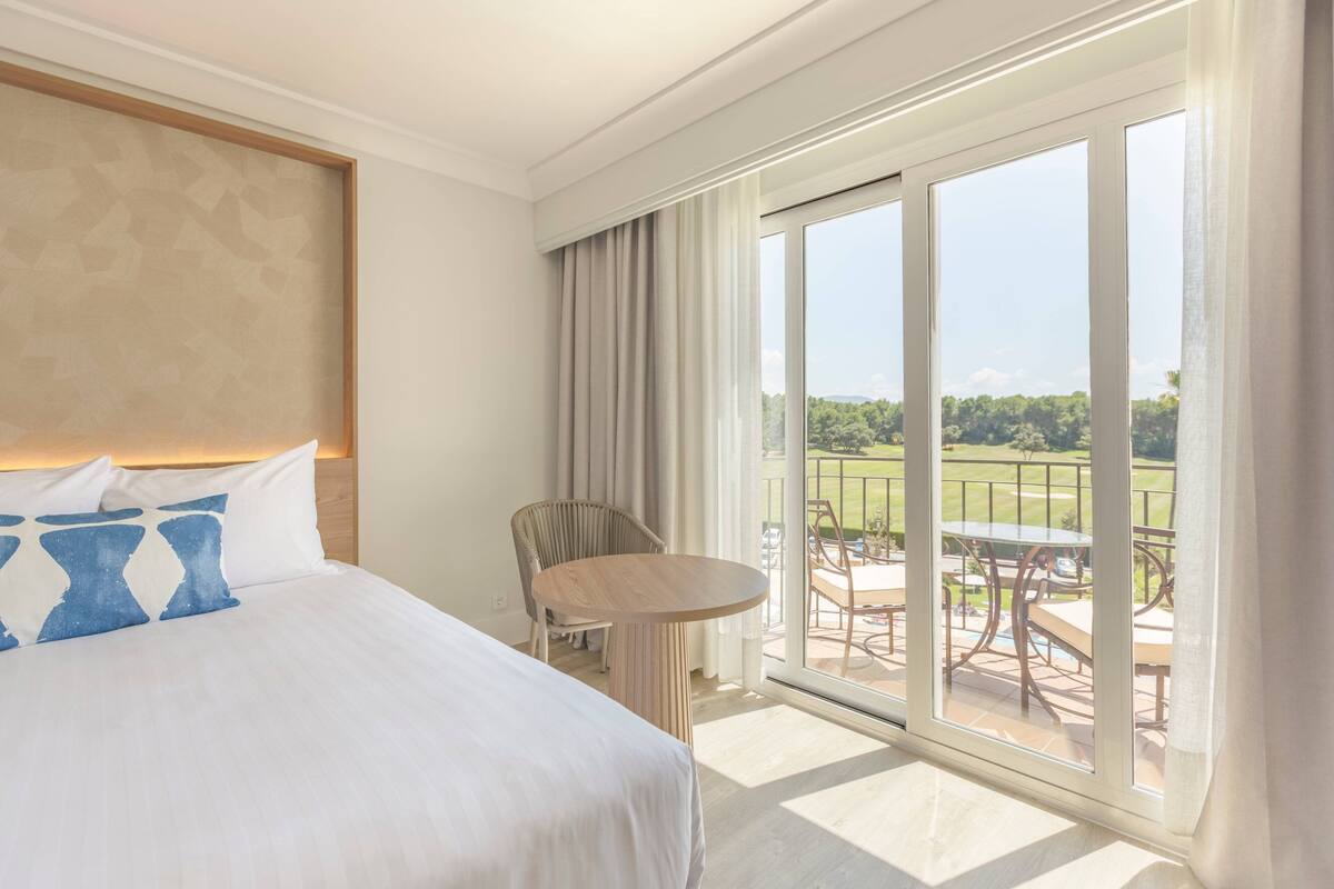 Quarto de hotel com uma cama grande, uma mesa redonda e uma varanda com vista para uma paisagem verde.