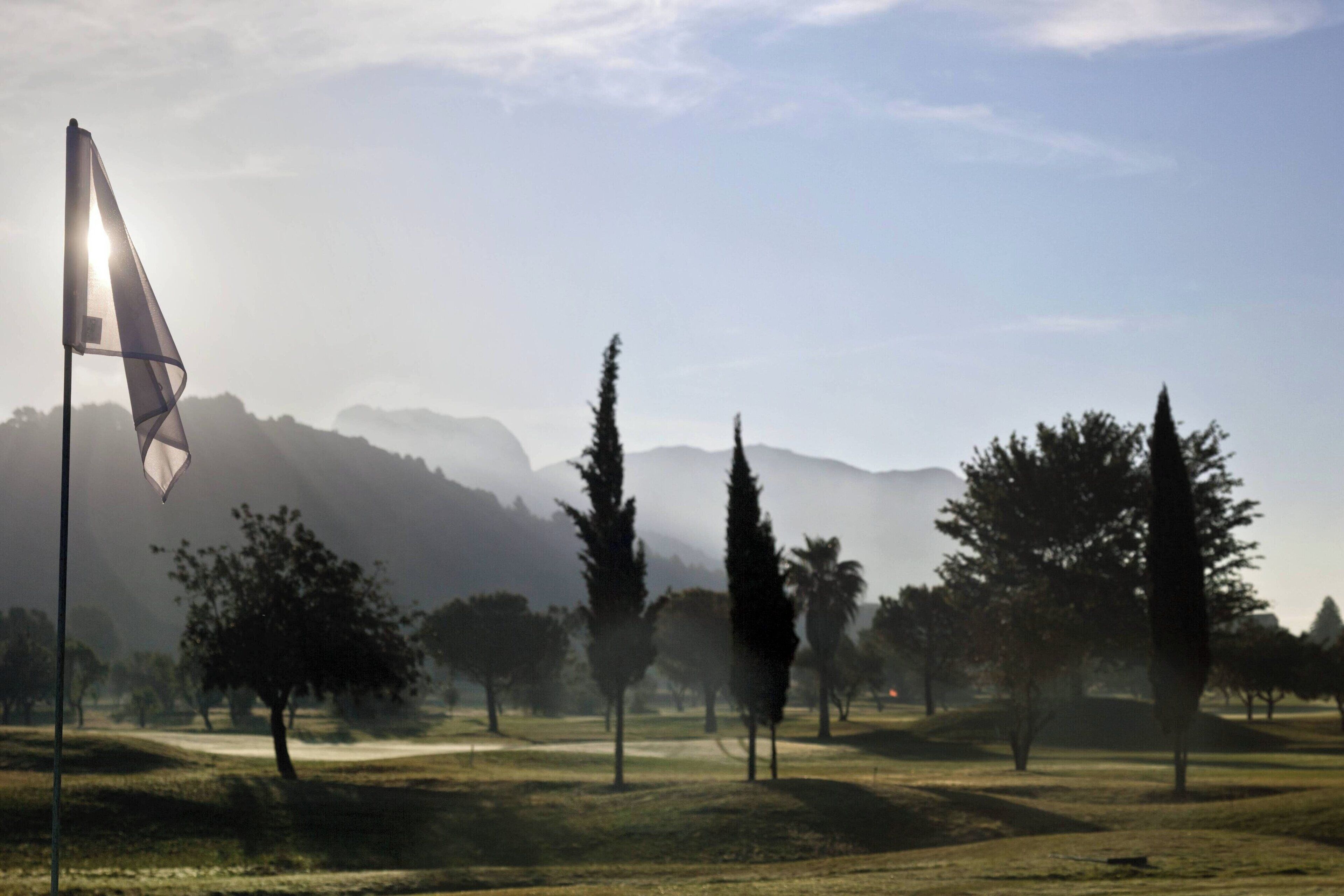 A golf course with a flag, trees, and mountains in the background.