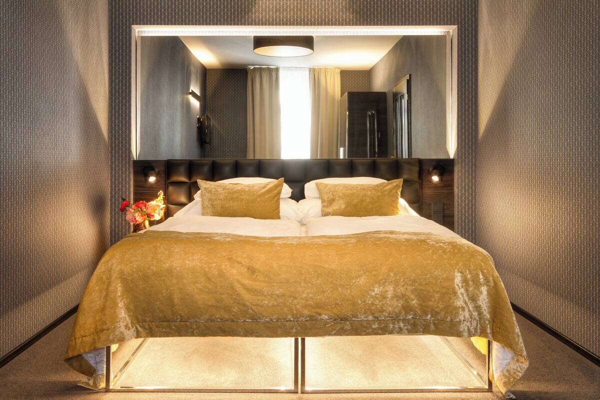 A modern hotel room with a large bed, a mirror above the bed, and a nightstand with a vase of flowers.