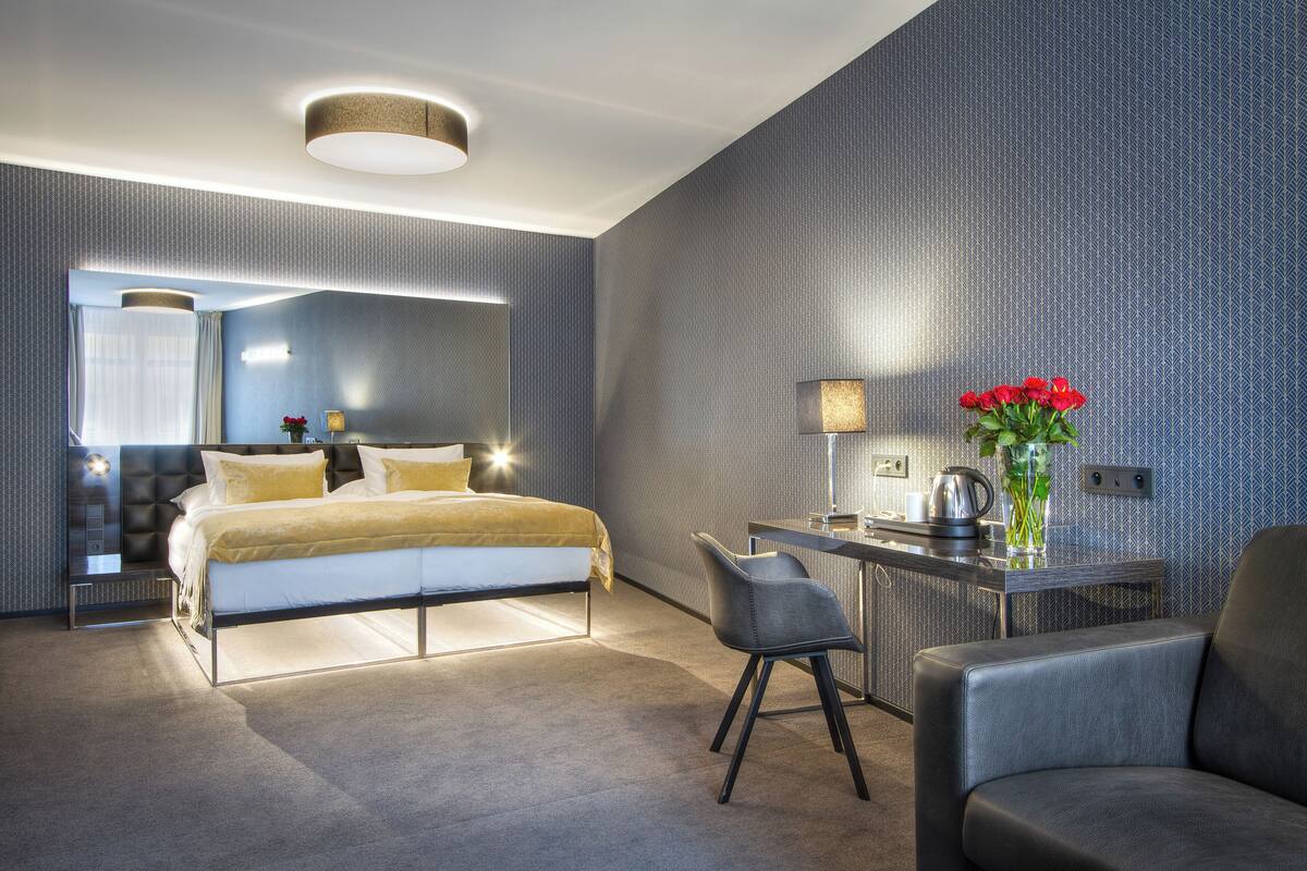 A modern hotel room with a bed, a desk, a chair, and a vase with red flowers.