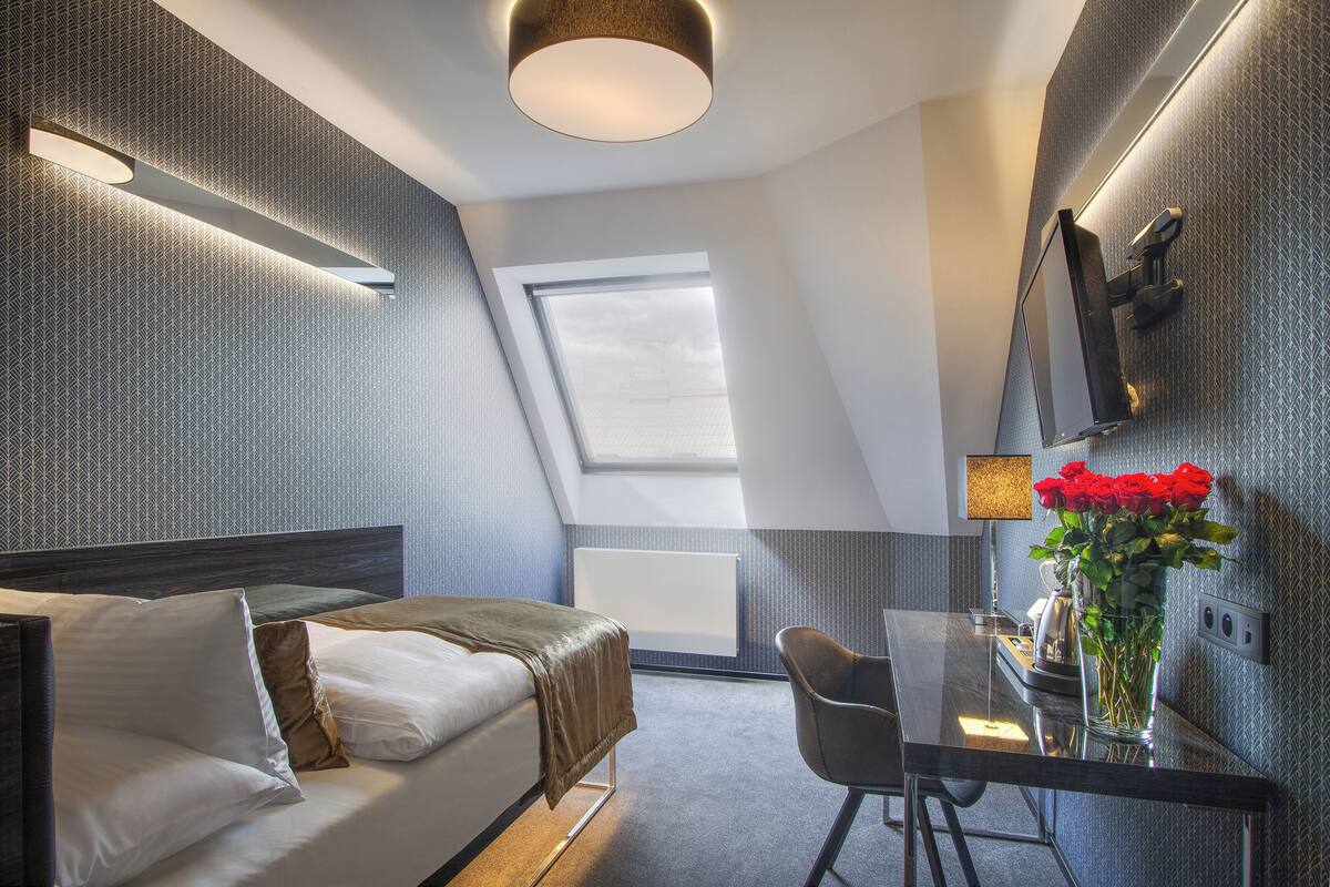 A modern hotel room with a bed, a desk, a chair, a large window, and a flat-screen TV mounted on the wall.