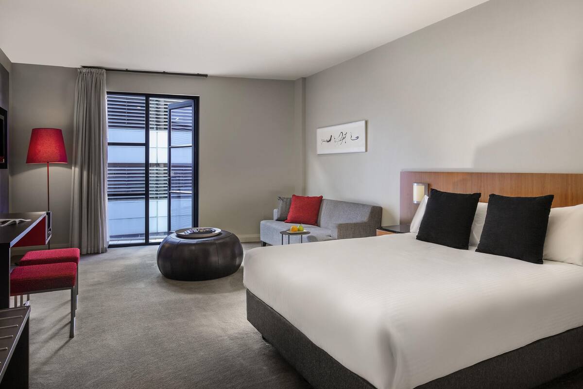 A hotel room with a bed, a desk, a chair, a round ottoman, a sofa, and a window with blinds.