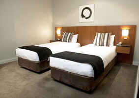 A hotel room with two beds, each with a wooden headboard and a black throw blanket. There are two bedside tables with lamps, and a wall-mounted artwork above the beds.
