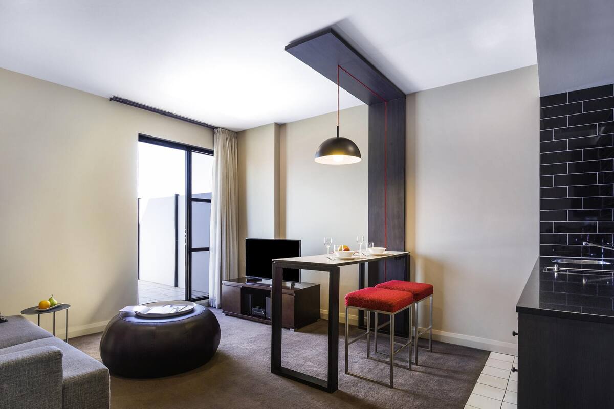 A modern hotel room with a sofa, a dining table with red stools, a TV, and a large window.