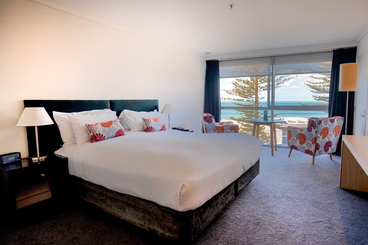A hotel room with a large bed, two chairs, a desk, and a view of the ocean.