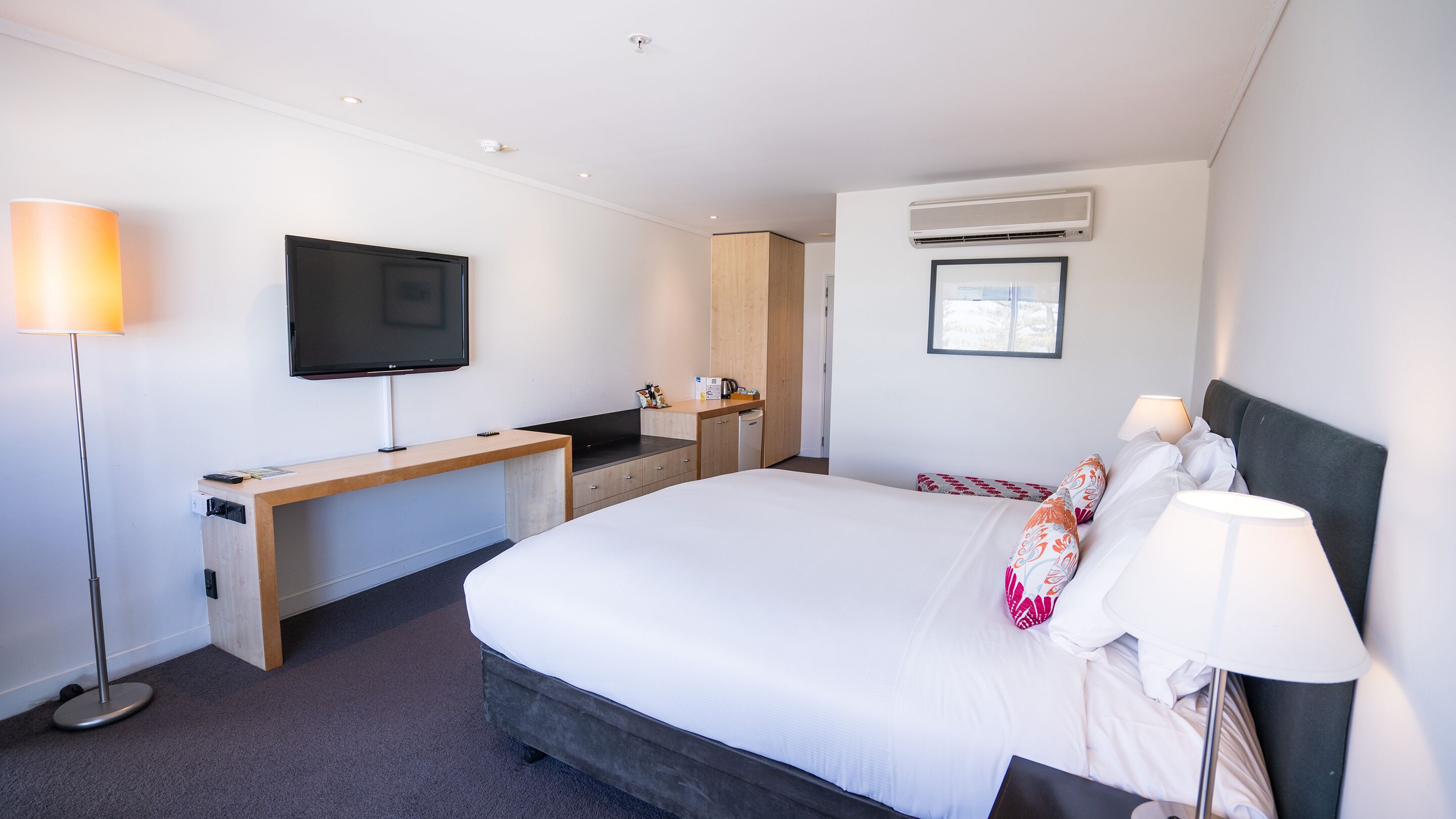 A modern hotel room with a large bed, a desk with a flat-screen TV, a built-in air conditioning unit, and a window providing natural light.